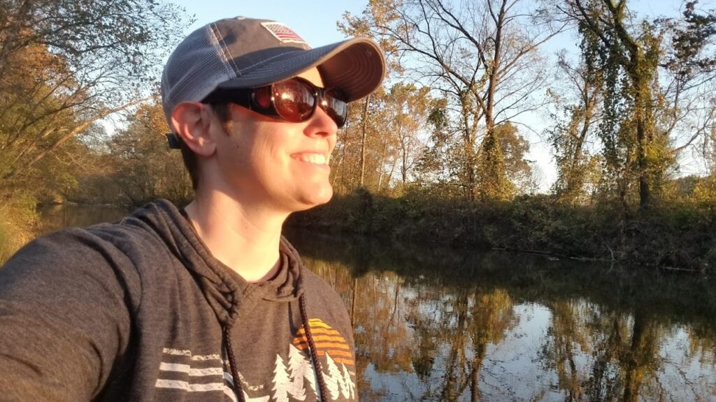 The author standing by a creek with a smile on her face, lit by a setting sun.