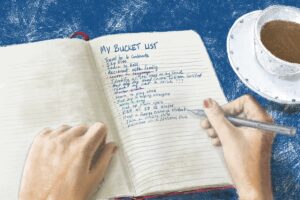A closeup of hands writing in a journal page titled "My Bucket List" with several items below it. Listen to S4E24 to learn about why it is good to have a bucket list