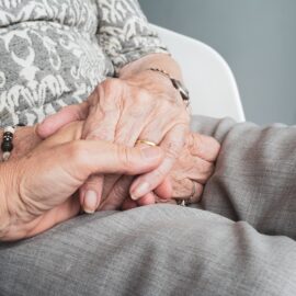 A younger person holding an elder’s hands. We talk about dementia and scams affecting seniors in this episode.