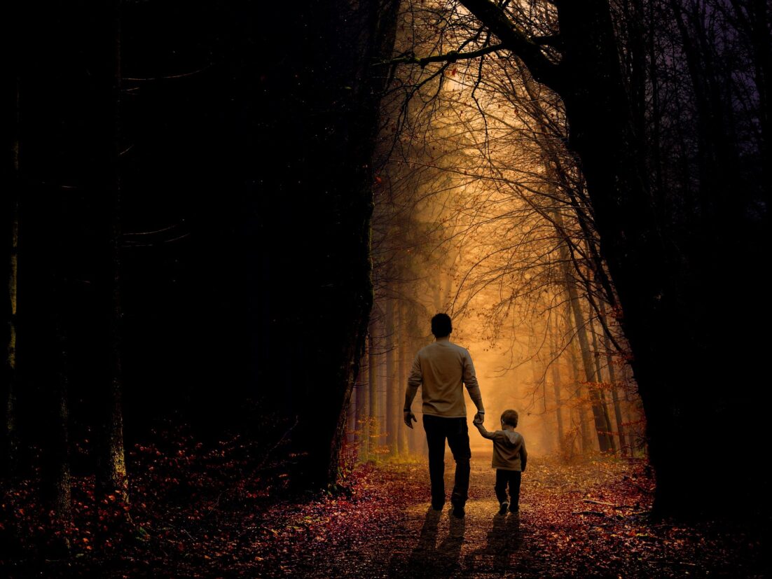 A father walking with his son, holding his hand through a dimly lighted forest. Learn how to talk to your children about death https://every1dies.org