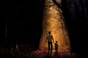 A father walking with his son, holding his hand through a dimly lighted forest. Learn how to talk to your children about death https://every1dies.org