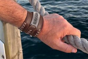 A close-up of a man's hand holding the rope railing by water. He has a leather bracelet with "survivor" on it