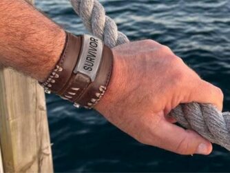 A close-up of a man's hand holding the rope railing by water. He has a leather bracelet with "survivor" on it