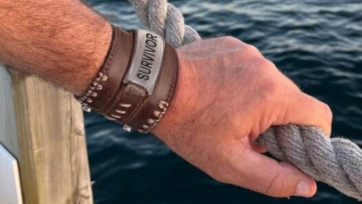 A close-up of a man's hand holding the rope railing by water. He has a leather bracelet with "survivor" on it
