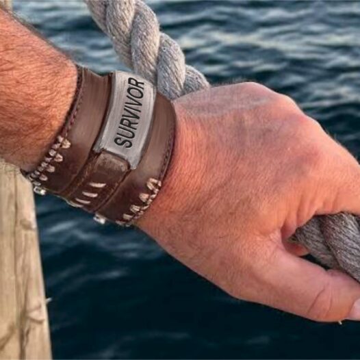 A close-up of a man's hand holding the rope railing by water. He has a leather bracelet with "survivor" on it