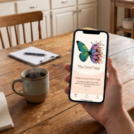 Hand holding a smartphone displaying The Grief App logo at a sunny kitchen table with a morning cup of coffee.