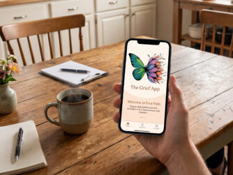 Hand holding a smartphone displaying The Grief App logo at a sunny kitchen table with a morning cup of coffee.